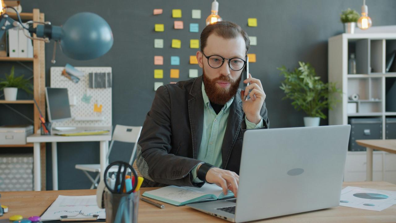 Pria di telepon bekerja di laptop di kantor